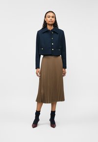 Granatowa krótka kurtka z złotymi guzikami, w połączeniu z brązową plisowaną midi spódnicą. Modelka ma na sobie czarne prążkowane skarpetki i czerwone szpilki.