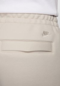 Gaiši beiges Nike sporta bikses ar strukturētu audumu, elastīgu jostasvietu un aizmugurējo kabatu ar smalku izšuvumu.