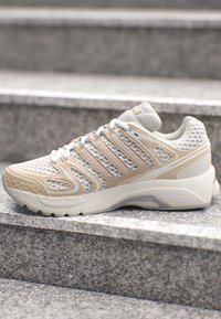Weißer und beiger Laufschuh mit Mesh- und synthetischen Overlays, der auf einer Außentreppe aus Stein liegt.