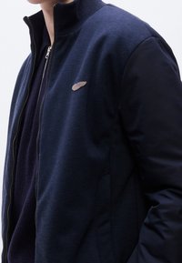 Personne portant une veste zippée bleu marine avec un petit emblème brodé en forme d’aile sur la poitrine gauche, superposée à un pull en maille foncé.