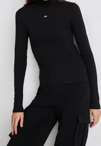 Schwarzer langärmliger Rollkragenpullover aus Rippmaterial mit kleinem Logo auf der Brust. Die Passform ist schlank und hat eine glatte Textur.