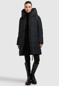 Sort pufferjakke med høj krave, frontknapper og snoretræk i hætten. Glat, quiltet stof. Modellen bærer sorte leggins og knæhøje støvler.