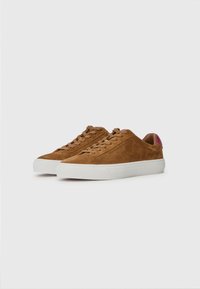 Zapatillas de ante marrón con suela de goma blanca, punta redonda y diseño de cordones. Pequeño acento rojo en el talón. Textura suave.