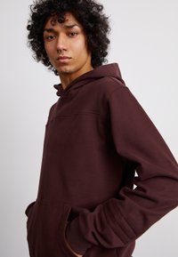 Hoodie bordeaux in morbido tessuto con tasca a marsupio, polsini testurizzati e vestibilità rilassata. Caratterizzato da un design minimalista e cappuccio con cordino.