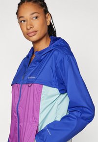Windbreaker jas in blauw, paars en lichtgroen. Beschikt over een capuchon, ritssluiting en verstelbare manchetten. Gemaakt van lichtgewicht, waterafstotend materiaal.