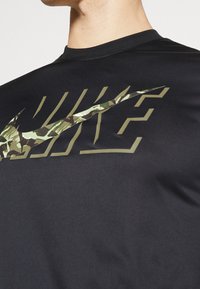 Camiseta deportiva negra con un gran logo de Nike en estampado de camuflaje verde, mangas cortas y cuello redondo. Tejido suave y ligero.