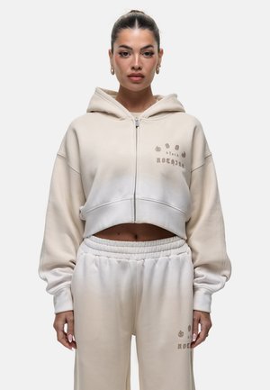 Beige Kapuzenpullover mit Reißverschluss, lockerer Schnitt und besticktem Text. Kombiniert mit passenden hochtaillierten Jogginghosen und elastischem Bund.