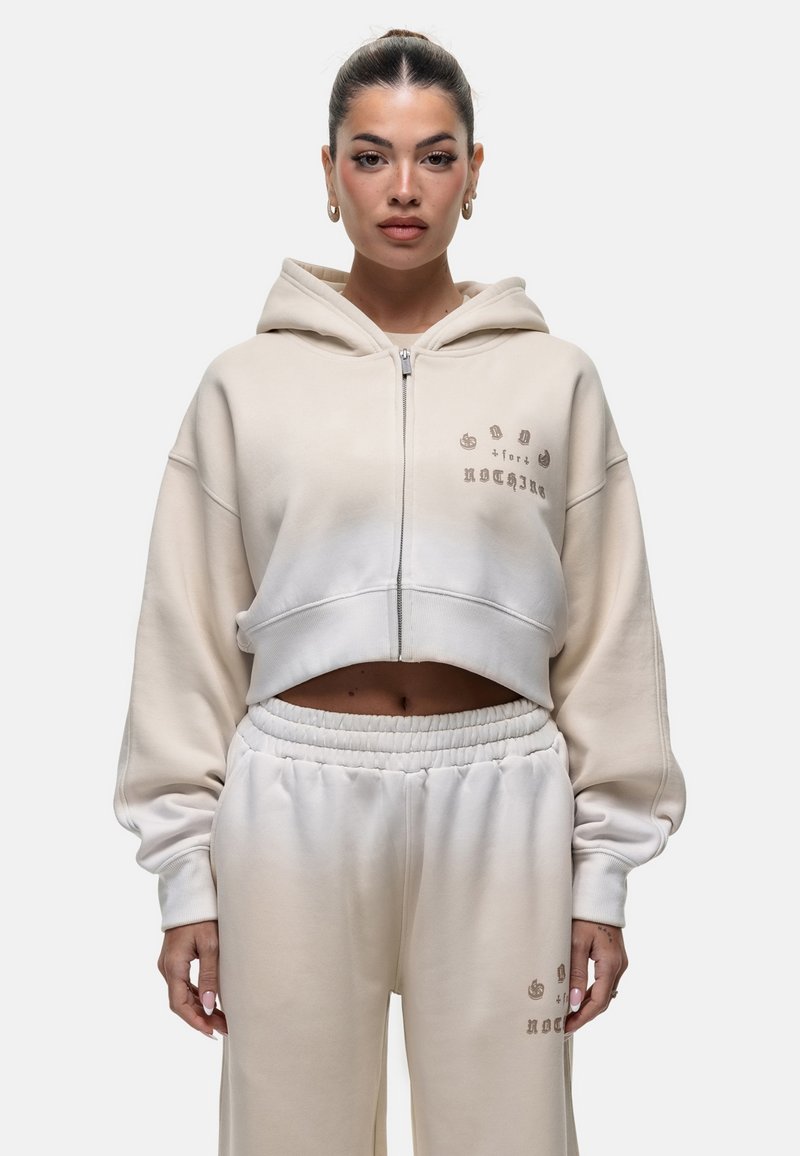 Haut à capuche beige coupé avec une fermeture éclair, coupe ample et texte brodé. Assorti à un pantalon de jogging taille haute avec ceinture élastique.