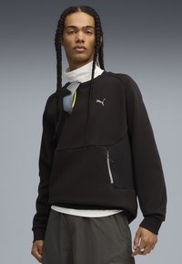 Felpa nera con un design testurizzato, tasca con zip e logo Puma grigio. Indossata sopra uno strato bianco con collo alto, abbinata a pantaloni corti grigi.