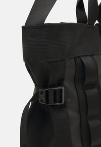 Czarna torba z materiału o teksturowej powierzchni, z regulowanymi paskami i plastikową klamrą. Posiada pionowy kształt oraz wzmocnione szwy.