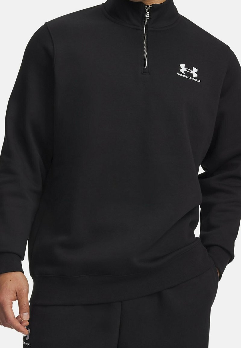 Haut noir à manches longues avec fermeture éclair jusqu'à la poitrine et logo Under Armour sur la poitrine, porté avec un pantalon noir assorti, mains partiellement visibles.