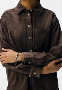 Brown corduroy shirt with a button-up front, long sleeves, and a chest pocket. Features textured vertical stripes and round buttons.