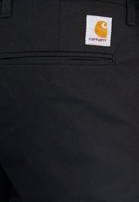 Black pants with a smooth texture, featuring a rear pocket and a white Carhartt logo tag with an orange accent. Simple and functional design.