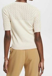 Beżowy sweter z krótkim rękawem, zdobiony misternymi wzorami dzianinowymi na ramionach i plecach, z prążkowanym dołem i gładką fakturą.