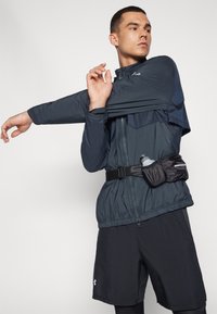 Athlète portant une veste sombre, un short et une ceinture avec une bouteille d'eau, étend son bras à travers sa poitrine sur un fond blanc.