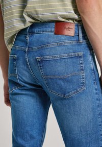 Blå jeans i denim med rak skärning, synliga sömmar och en brun läderlapp vid midjan. Har två olika bakficksstilar.