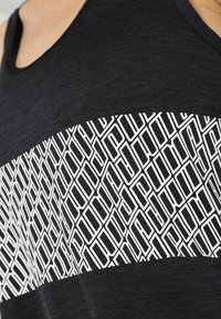 Černý top na ramínka s texturovanou tkaninou a velkým bílým geometrickým vzorem přes hruď. Jednoduchý design bez ozdob.