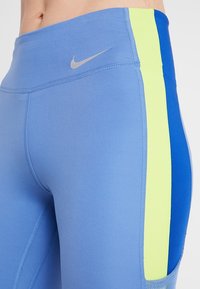 Detailní záběr osoby v modrých sportovních legínách se žlutými a tmavě modrými bočními pruhy a malým šedým logem Nike na pase.