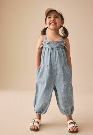 Smilende pige med fletninger iført en lyseblå blomsterprintet jumpsuit, beige kasket og hvide sandaler med spænde, stående mod en ensfarvet baggrund.