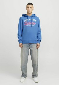 Sudadera con capucha azul y estampado frontal, con puños y dobladillo de canalé. Combinada con vaqueros de un azul claro y zapatillas blancas. Diseño sencillo.