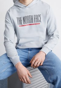 Persona vistiendo una sudadera gris clara con el texto "THE NORTH FACE" y el eslogan rojo "NEVER STOP EXPLORING", combinada con jeans azules, sentado en un banco metálico.