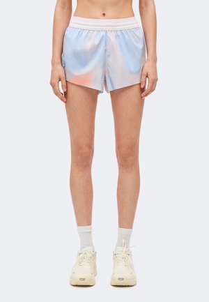 Persona che indossa pantaloncini tie-dye azzurro chiaro e rosa con fascia bianca in vita, calzini bianchi e sneaker color panna, in piedi davanti a uno sfondo semplice.