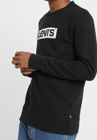 Sudadera negra con cuello redondo, que presenta un prominente logo blanco de "Levi's" en el frente y puños y dobladillo acanalados. Diseño minimalista.