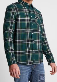 Vihreä ruutukuosisella napitettavalla paidalla, joka on valmistettu kudotusta kankaasta; siinä on valkoisia ja mustia viivoja, rintatasku ja pitkät hihat.