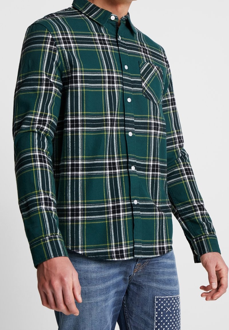 Vihreä ruutukuosisella napitettavalla paidalla, joka on valmistettu kudotusta kankaasta; siinä on valkoisia ja mustia viivoja, rintatasku ja pitkät hihat.