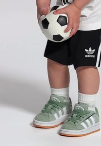 Zelené semišové tenisky s bílými pruhy, oranžovou gumovou podešví a tkaničkami. Dítě drží černo-bílý fotbalový míč a má na sobě černé kraťasy.