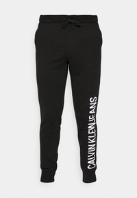 Černé tepláky vyrobené z bavlny, s elastickým pasem na šňůrku a zúženými kotníky, se velkým bílým textem "CALVIN KLEIN JEANS" na boku.