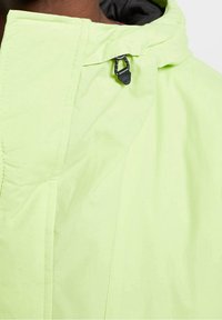 Primer plano de una persona que lleva una chaqueta con capucha de color verde claro, con un tirador de cremallera negro visible cerca del cuello.