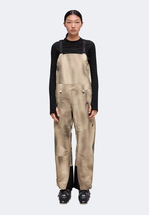 Personne portant une salopette beige avec un t-shirt noir à manches longues et des bottes de neige noires, debout devant un fond clair uni.