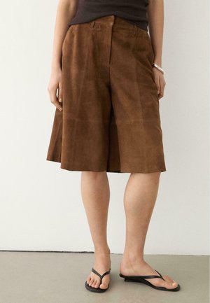 Person iført brune knælange ruskindsshorts, sort ærmeløs top, sølvarmbånd og sorte thongsandaler, stående på grå gulv.