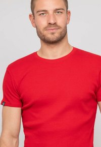 T-shirt rouge à côtes avec des manches courtes, un col rond et un petit logo sur la manche. Le tissu semble doux avec une texture lisse.