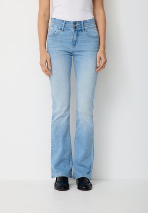 Frau trägt hellblaue, hoch taillierte Schlagjeans, weißes Oberteil und schwarze Loafers, steht vor einem schlichten weißen Hintergrund.