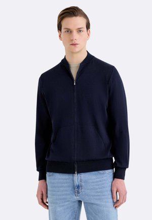Giovane uomo che indossa una giacca blu navy con zip sopra una camicia beige e jeans azzurri, in piedi davanti a uno sfondo chiaro e uniforme.