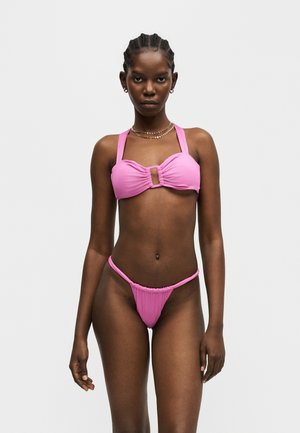 Kobieta z zaplecionymi włosami, ubrana w różowy prążkowany bikini z kwadratowym wycięciem na górze, stojąca na jednolitym jasnoszarym tle.