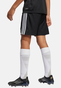 Camiseta y pantalones cortos negros con tres rayas blancas, calcetas blancas con detalles de malla y botas de fútbol negras con acentos plateados.