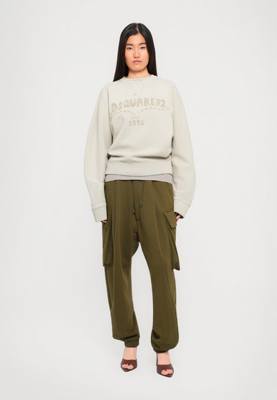 Young woman wearing beige oversized sweatshirt with "DSQUARED2 Since 1995" and olive green cargo pants with pockets, standing against white background.