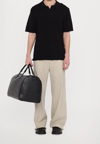 Mies, joka on pukeutunut mustaan lyhytnokkaiseen polo-paitaan ja beigeihin housuihin, pitää mustaa nahkaduffelilaukku k left kädessä.