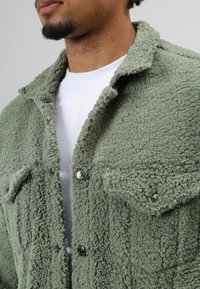 Green fuzzy jacket made of textured material, featuring a buttoned front, two chest pockets, and a collared neckline.