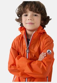 Chaqueta impermeable naranja con cremallera frontal, capucha elástica y detalles en rojo y blanco; presenta un parche con logo en el brazo izquierdo.