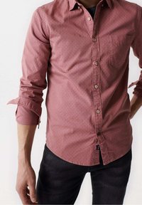 Camicia a maniche lunghe, con bottoni, di colore rosa polveroso con un motivo a pois discreto, dotata di tasca e maniche arrotolate. Indossata con jeans scuri.