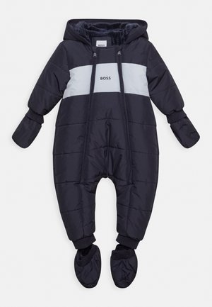 Marineblauwe gewatteerde babypak voor in de sneeuw met een grijze horizontale streep en logo. Heeft een capuchon, elastische manchetten en aangehechte voetjes. Zachte binnenvoering.