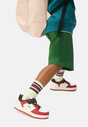 Child wearing red and white sneakers, striped socks, green shorts, teal sweatshirt, and carrying a light pink backpack mid-step.