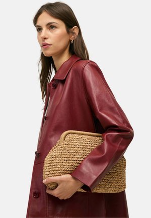 Mujer joven con abrigo de cuero rojo oscuro sosteniendo un gran bolso de mano beige tejido sobre un fondo liso.