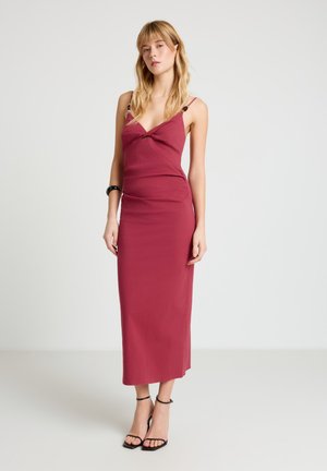 Donna con capelli biondi che indossa un vestito senza maniche, lungo fino alle caviglie, di colore rosa scuro con spalline sottili e sandali neri con tacco alto, in piedi davanti a uno sfondo semplice.