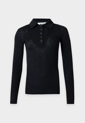Femme debout, main dans la poche, portant un polo noir à manches longues, un jean large bleu et des bottines noires sur un fond uni.