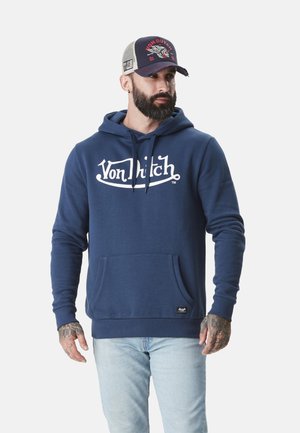 Homme barbu et tatoué portant un sweat à capuche Von Dutch bleu marine, un jean bleu clair et une casquette Von Dutch bleu marine et grise, regardant vers la droite.